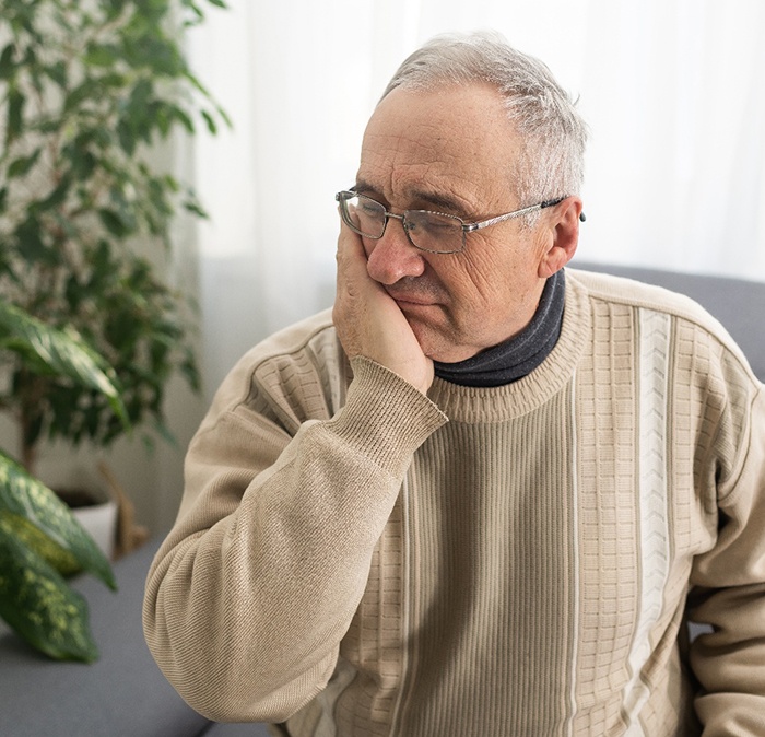 Senior man in sweater sitting on couch and rubbing jaw