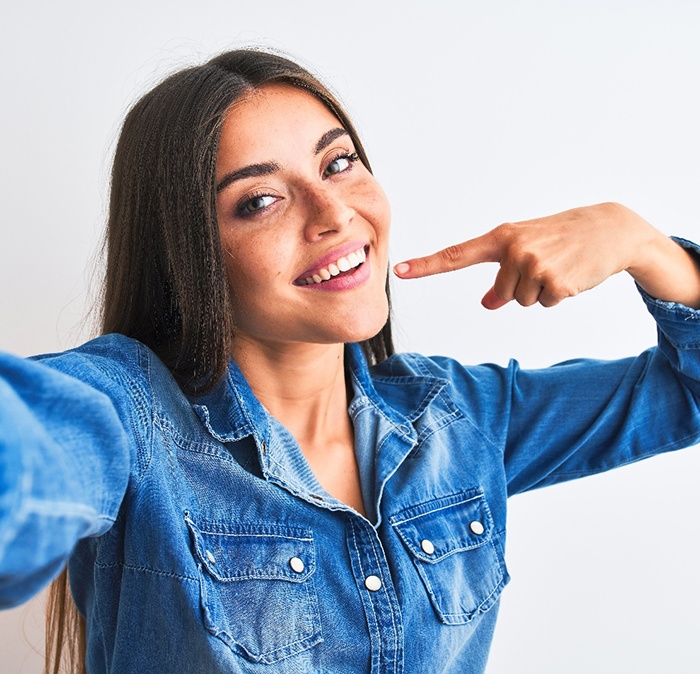 Woman in denim shirt taking selfie while pointing to her smile